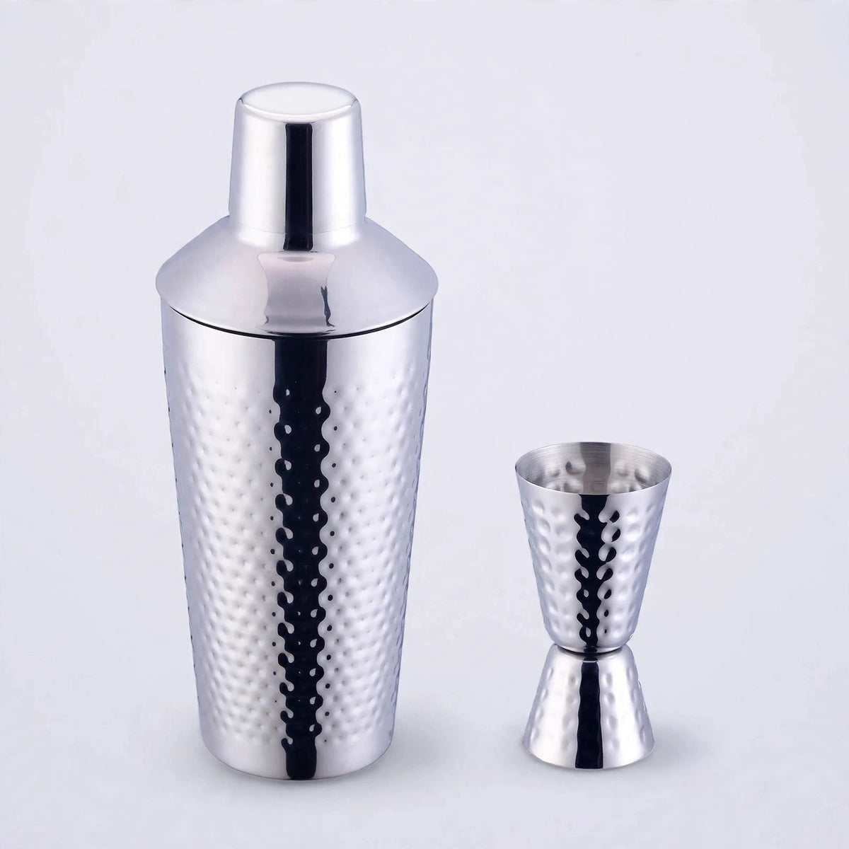 Silver hammered stainless steel cocktail shaker with matching double jigger on a plain light studio background.