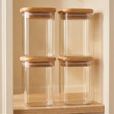 Four empty clear glass jars with bamboo lids are stacked in two columns on a wooden shelf against a beige background.