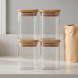 Four empty clear glass jars with bamboo lids stand stacked in two columns on a white counter beside a brown paper bag.