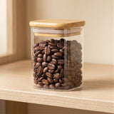 A clear glass jar filled with brown coffee beans and topped with a bamboo lid sits on a wooden shelf beside a window.