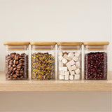 Four clear glass jars with bamboo lids hold coffee beans, dried plant pieces, white cubes, and red beans on a wooden shelf.