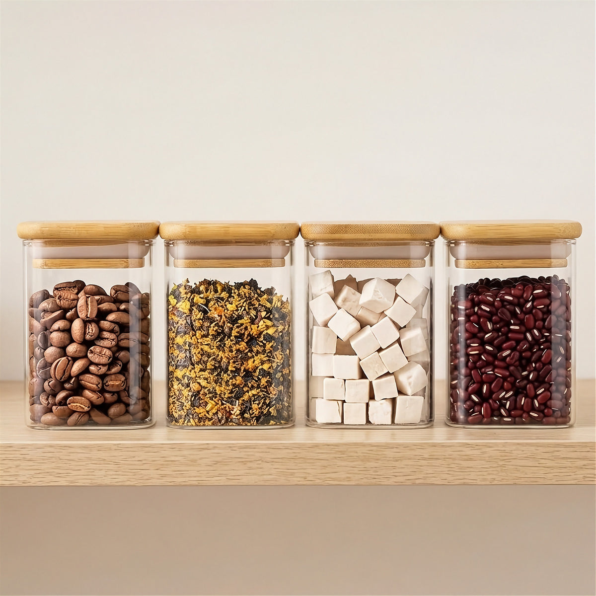 Four clear glass jars with bamboo lids hold coffee beans, dried plant pieces, white cubes, and red beans on a wooden shelf.