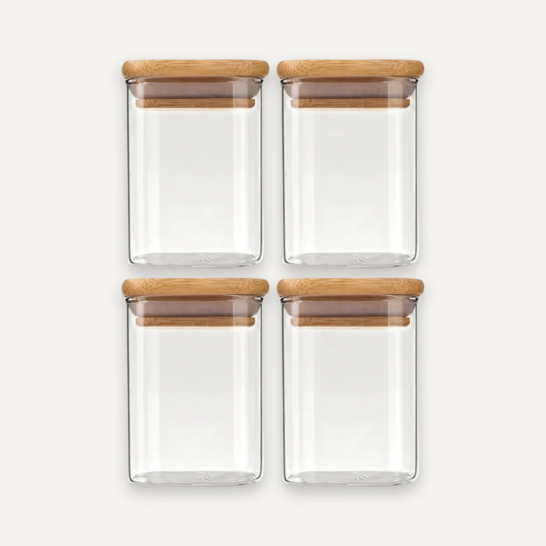 Four empty clear glass jars with bamboo lids are arranged in two rows of two against an off-white background.