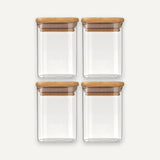 Four empty clear glass jars with bamboo lids are arranged in two rows of two against an off-white background.