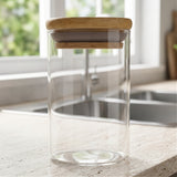 An empty clear glass jar with a bamboo lid stands on a light countertop beside a kitchen sink and window.