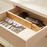 A natural bamboo drawer divider separates silver cutlery from dark cooking utensils in an open cream kitchen drawer.