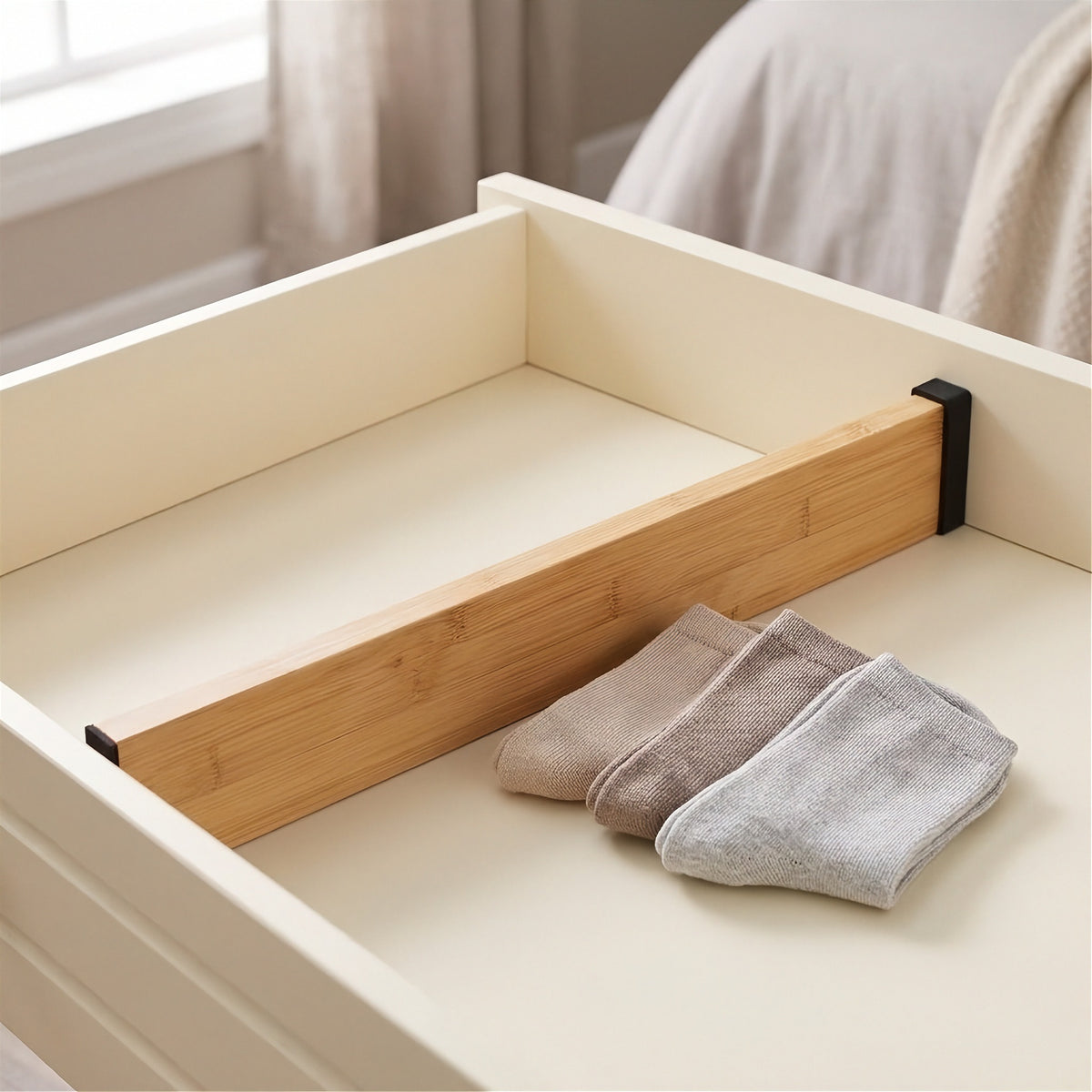 A light brown bamboo divider with black end pads spans a cream drawer beside three folded pairs of beige and gray socks.