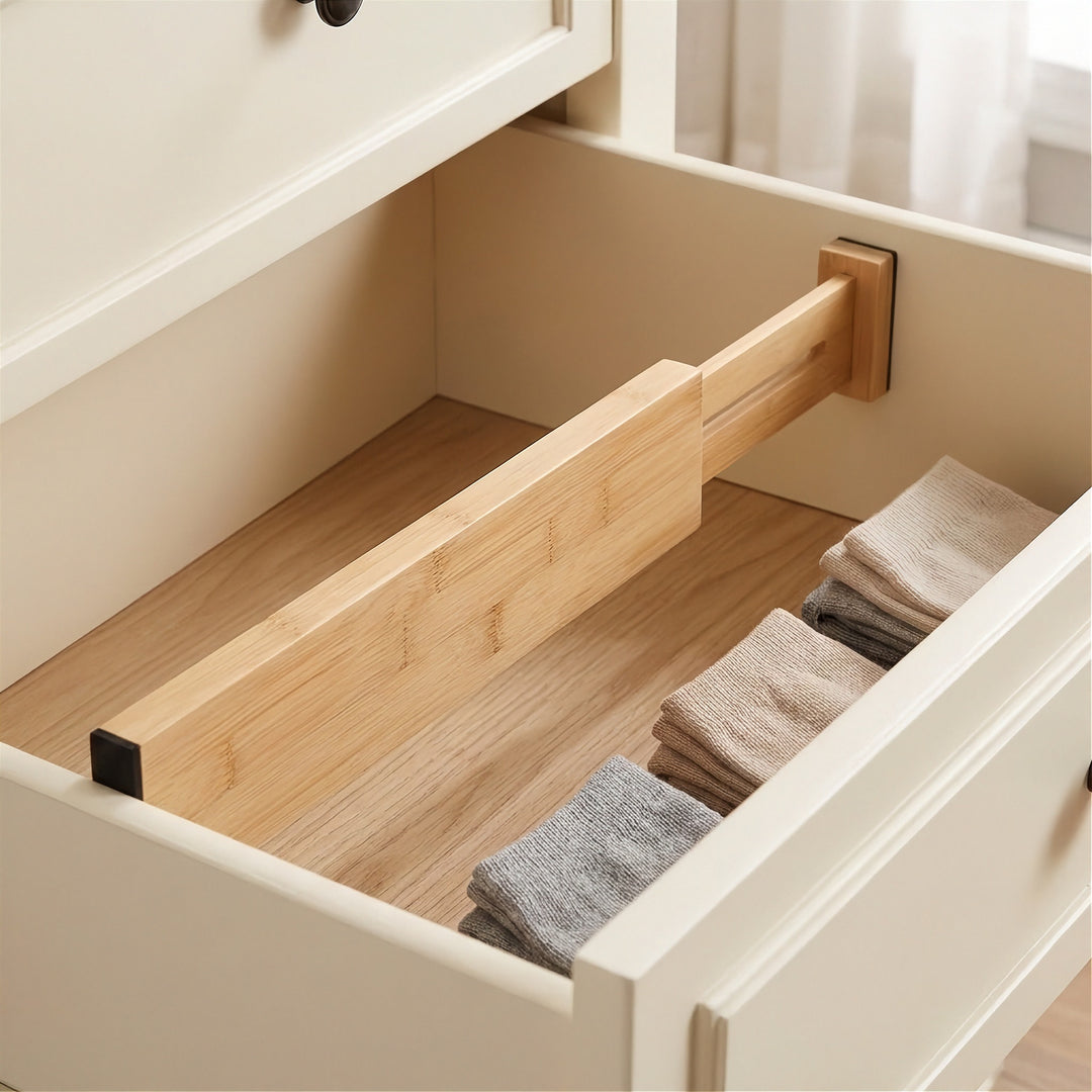 A light brown bamboo divider separates empty space from folded beige and gray socks inside a cream dresser drawer with a wooden base.