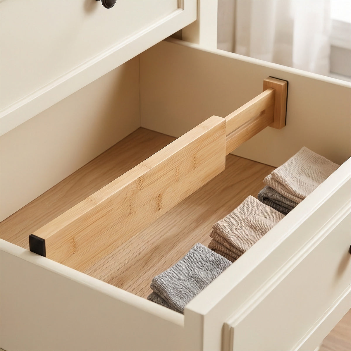 A light brown bamboo divider separates empty space from folded beige and gray socks inside a cream dresser drawer with a wooden base.