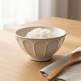 A ceramic bowl filled with white rice sits on a light wood table, with a wooden spoon resting on a folded beige cloth nearby.