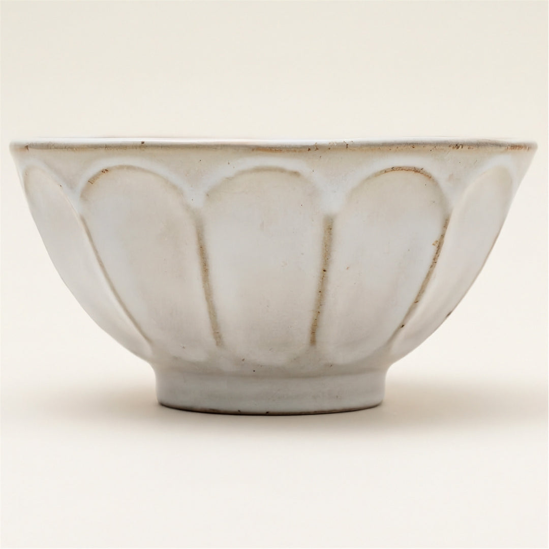 A white ceramic bowl with a scalloped, lotus-like exterior pattern and a small pedestal base, photographed against a solid off-white background.