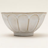 A white ceramic bowl with a scalloped, lotus-like exterior pattern and a small pedestal base, photographed against a solid off-white background.