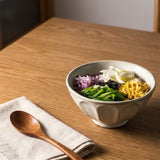 A ceramic bowl filled with colorful food sits on a wooden table next to a wooden spoon on a folded cloth napkin.