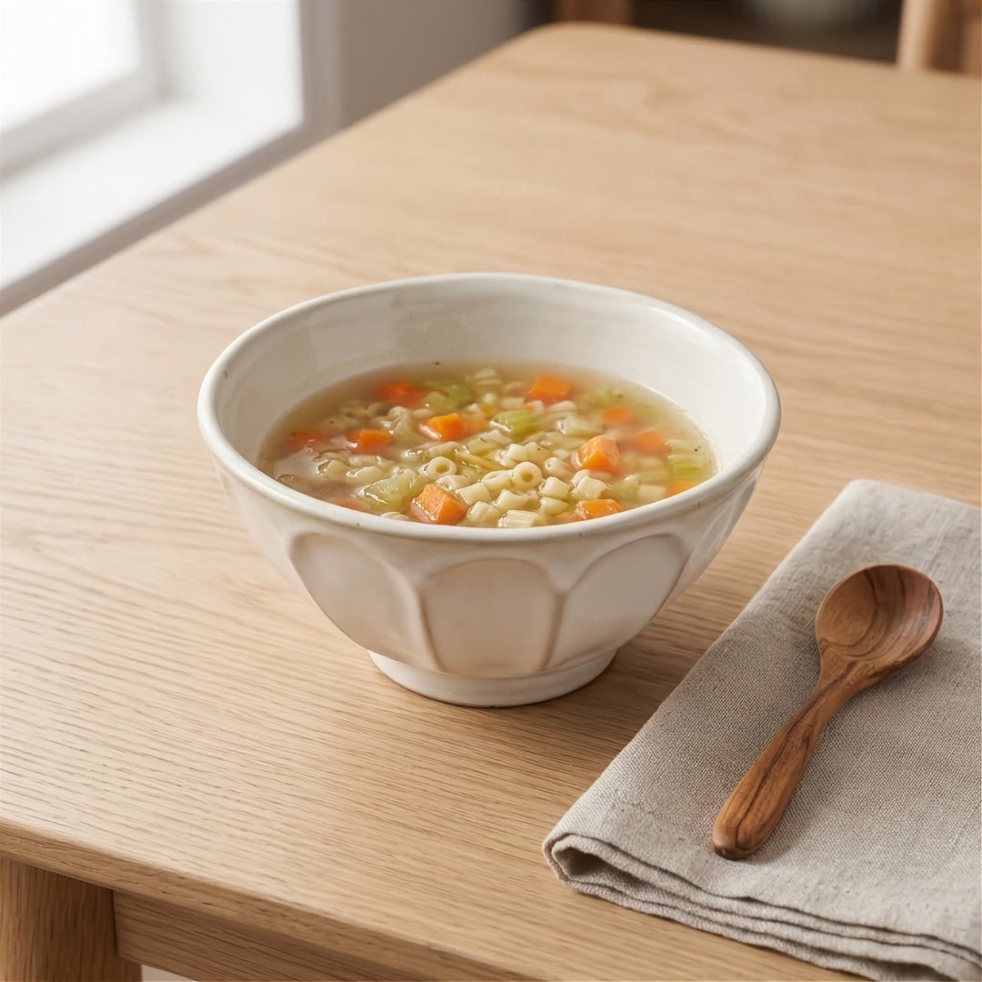 A ceramic bowl filled with a clear soup containing small pasta-like pieces and orange carrot chunks, placed on a light wood table beside a folded beige cloth and a wooden spoon.