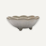 A small ceramic flower-shaped dish with three short feet, featuring a matte off-white glaze with subtle brown speckling along the rim, set against a solid off-white background.