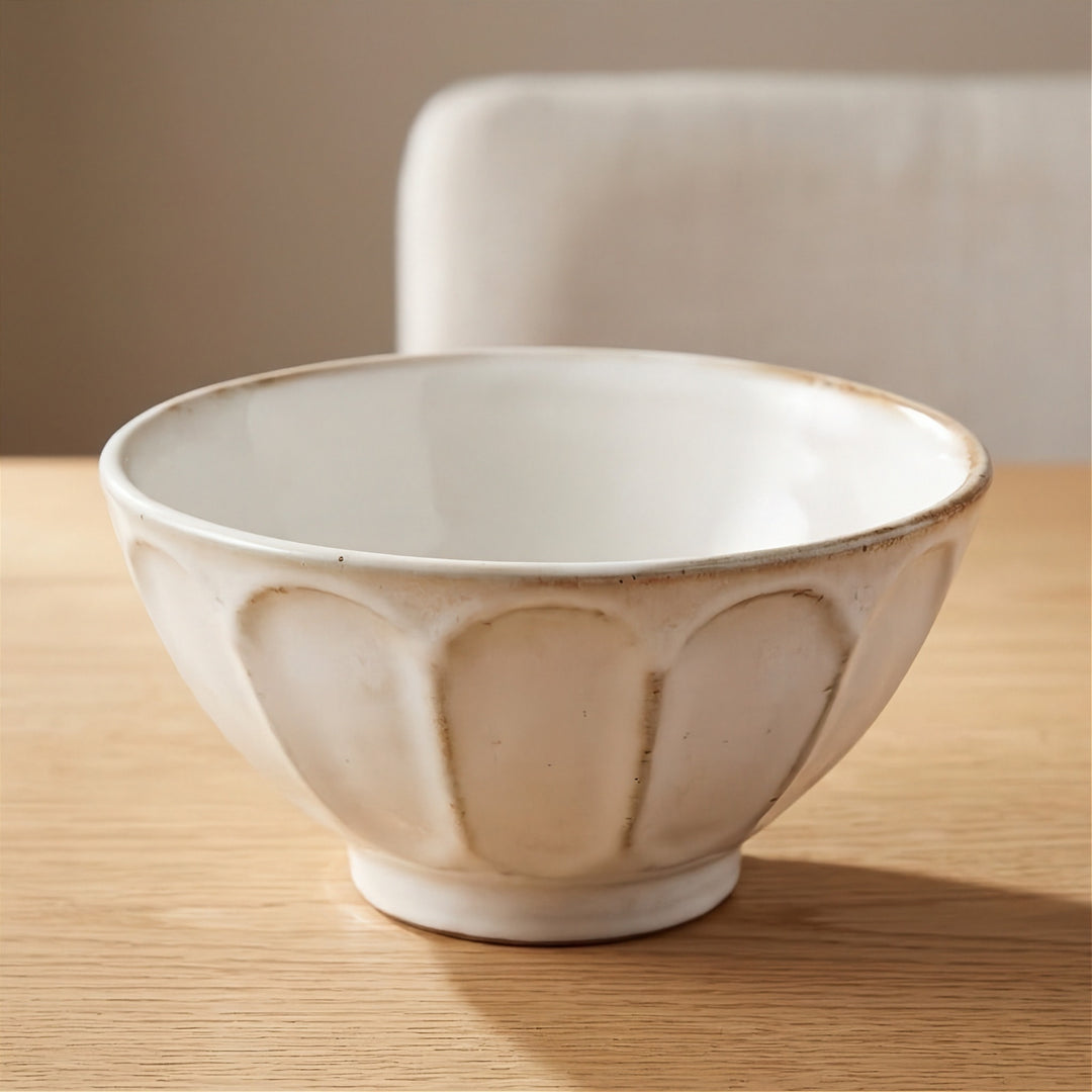 A ceramic bowl with a textured, matte off-white finish and a scalloped petal-like pattern on the exterior, sitting on a light wooden table with a blurred neutral background.
