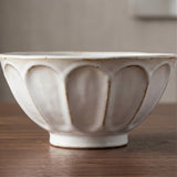 A ceramic bowl with a textured, ribbed exterior and a rustic, hand-painted finish, sitting on a wooden tabletop against a soft-focus neutral background.