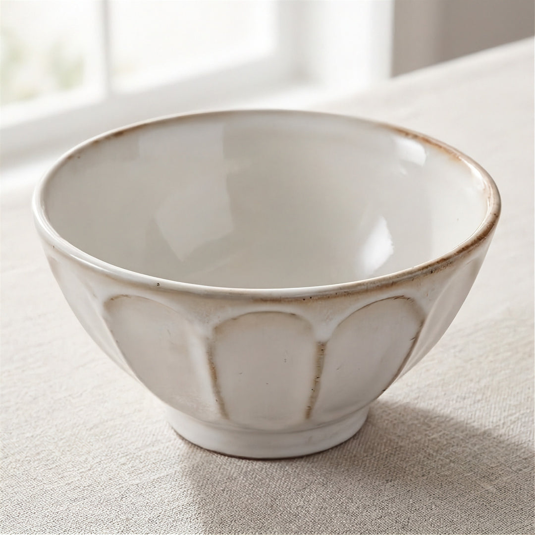 A white ceramic bowl with a textured, scalloped exterior and a rustic brown rim, resting on a beige linen tablecloth in a softly lit room.