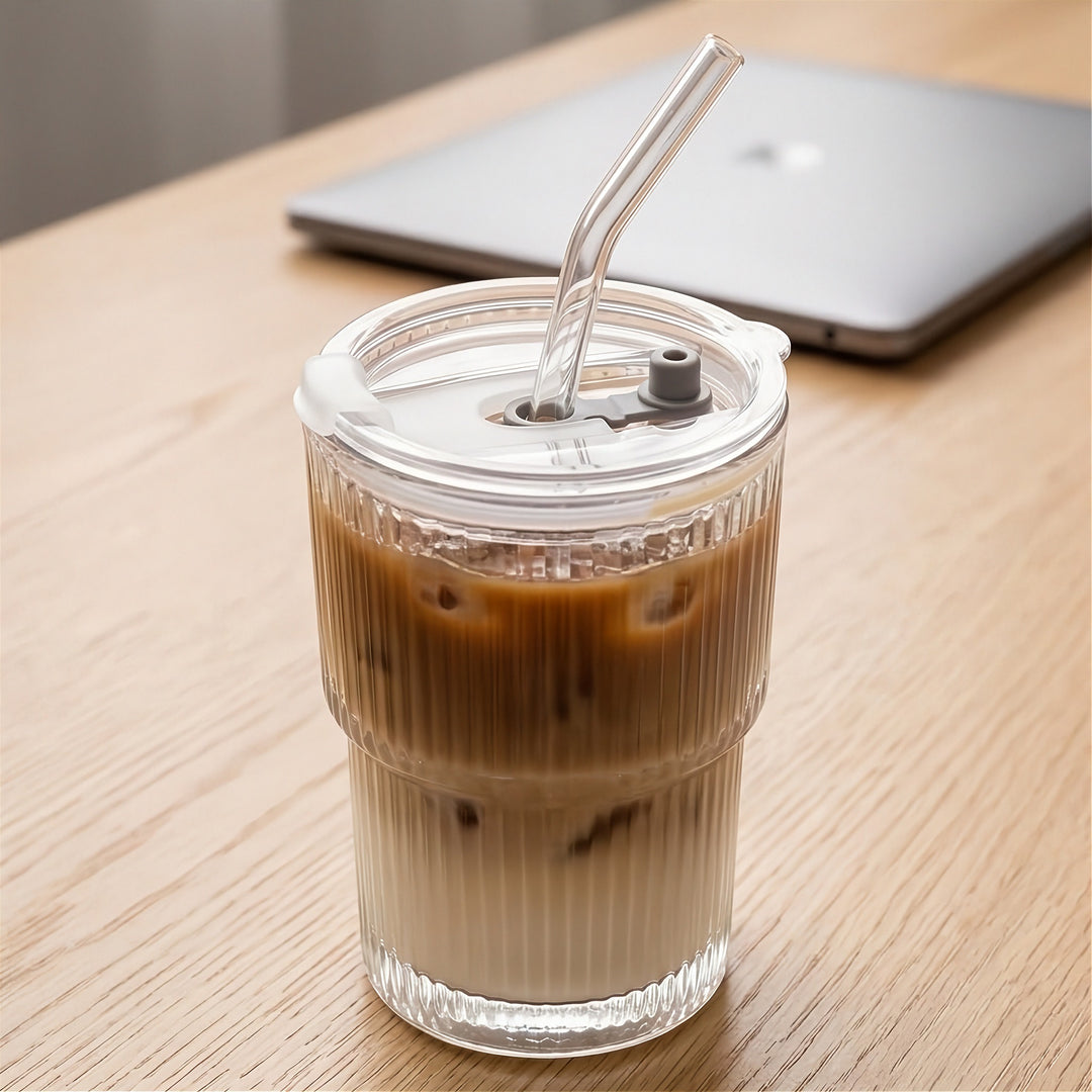 Clear ribbed tumbler with a transparent lid and bent straw holds layered iced coffee on a wooden desk beside a closed laptop.