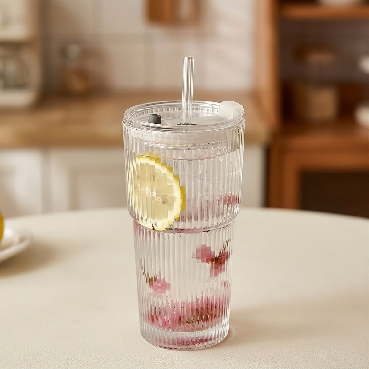 Clear ribbed tumbler with a transparent lid and straight straw holds water, a yellow lemon slice, and pink petals on a cream table.