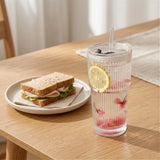 Clear ribbed tumbler with a lid and straw holds water, a lemon slice, and red fruit beside a sandwich on a wooden table.