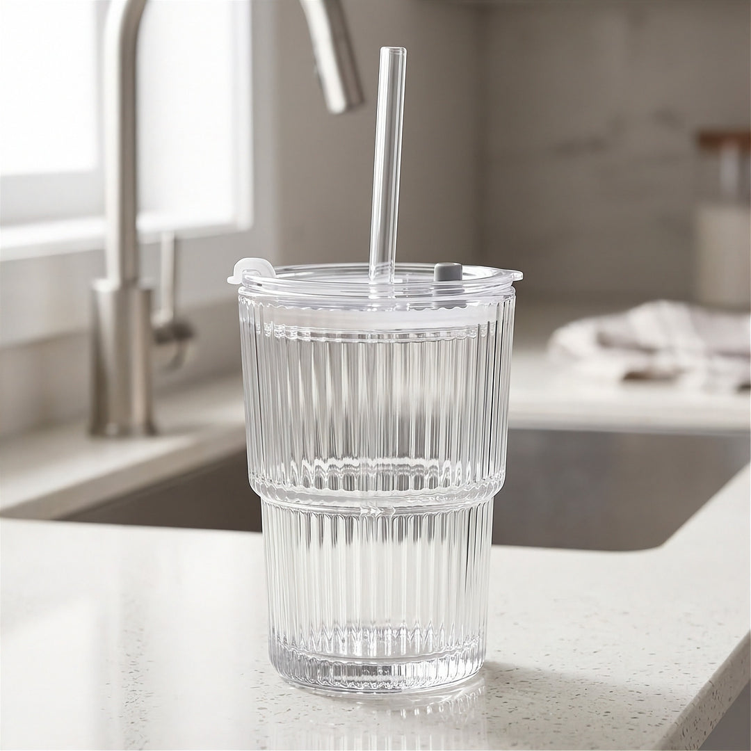 Empty clear ribbed tumbler with a transparent lid and straight straw stands on a light kitchen counter beside a sink and faucet.