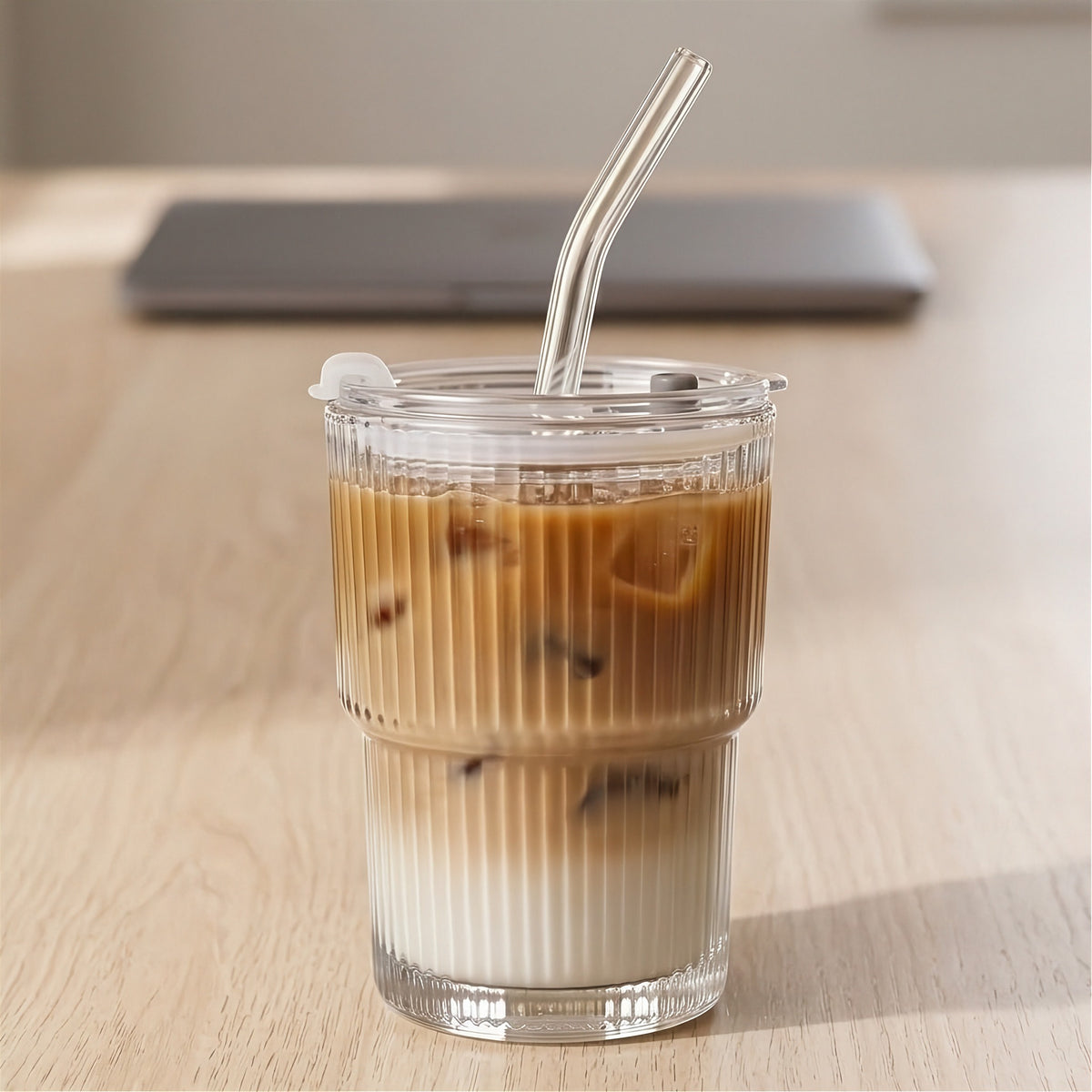 Clear ribbed tumbler holding a layered brown and white iced drink has a transparent lid and bent clear straw on a wooden table.