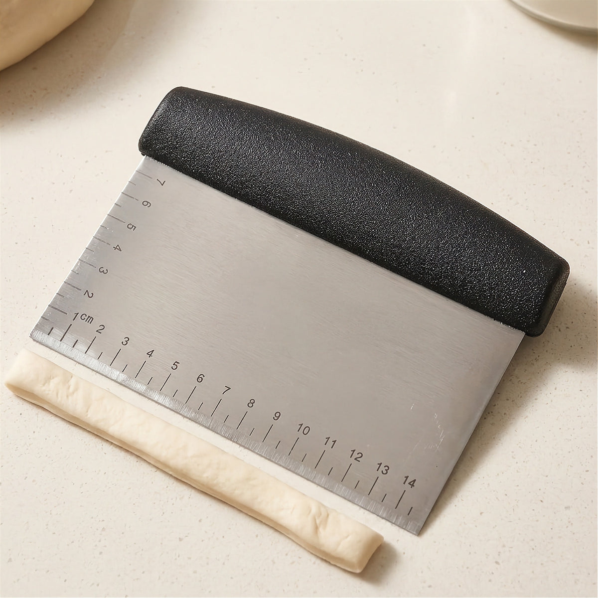 Bench scraper with etched centimeter markings measuring a rolled dough log on a light countertop