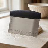 Stainless steel scraper standing upright on a floured countertop beside a rising dough ball