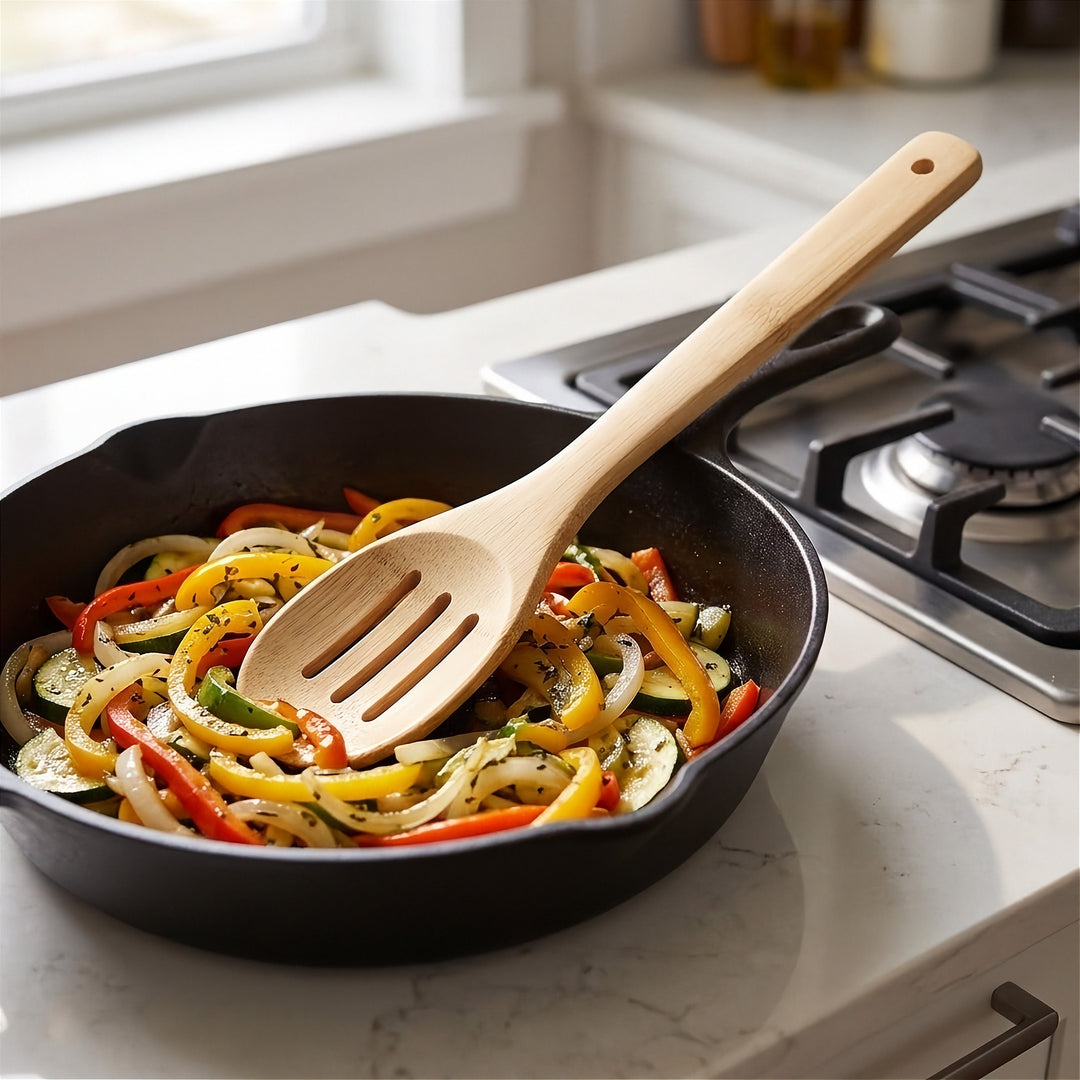 A natural light wood kitchen utensil with a slotted head resting in a black cast iron skillet filled with sautéed vegetables, set on a white marble kitchen countertop next to a gas stovetop.