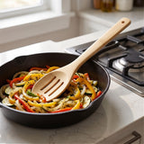 A natural light wood kitchen utensil with a slotted head resting in a black cast iron skillet filled with sautéed vegetables, set on a white marble kitchen countertop next to a gas stovetop.