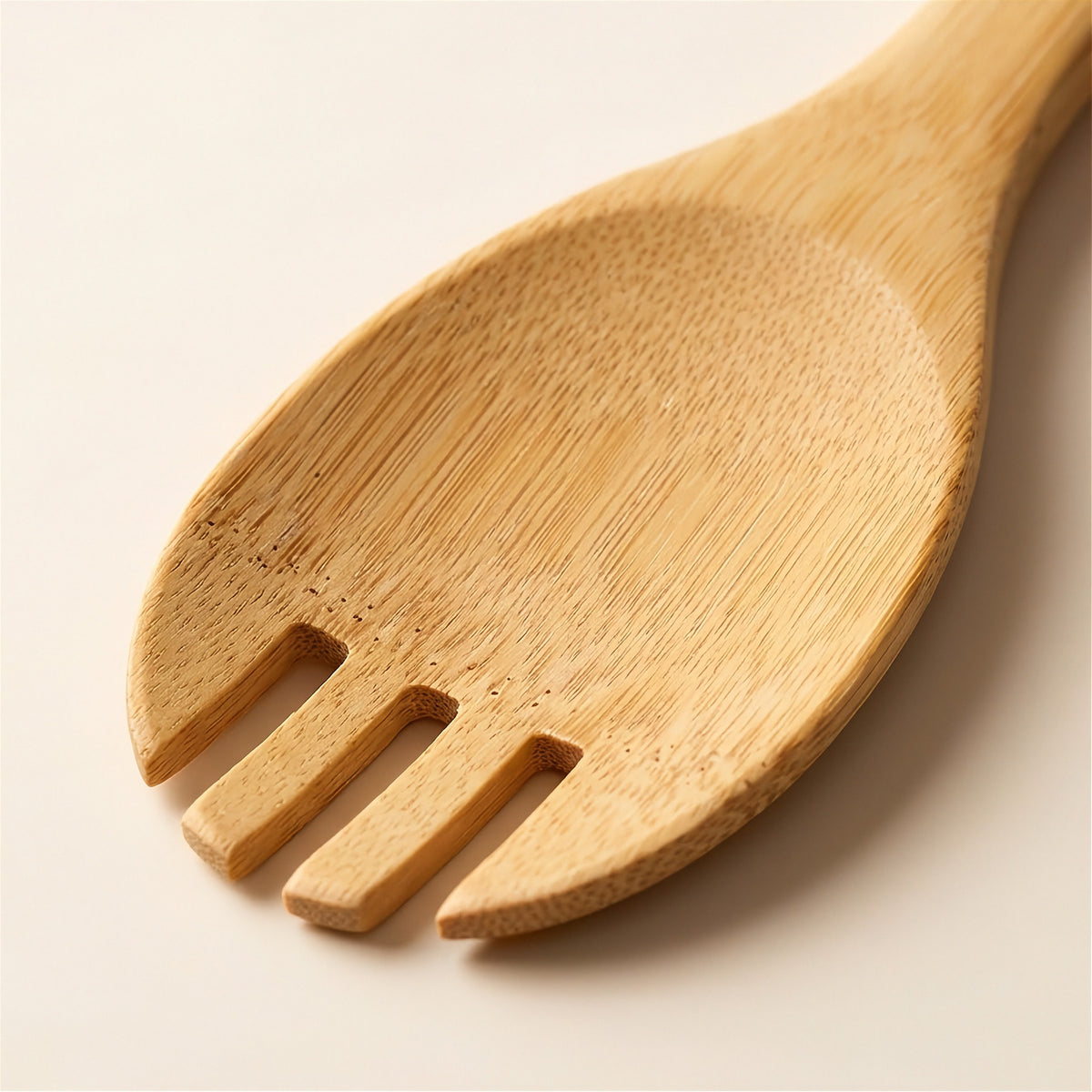 A close-up of a light natural bamboo serving spoon or salad server with three tines, showing the wood grain texture on a warm off-white background.