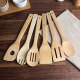 Six bamboo kitchen utensils, including a spoon, fork, spatula, and two slotted spoons, arranged on a dark wood table next to a beige linen cloth.