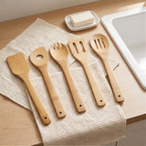 Five light natural bamboo kitchen utensils with wooden handles are arranged on a textured cream dish towel on a light wood countertop, with a white sink and a bar of soap visible in the background.