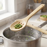 A bamboo kitchen utensil with a slotted bowl is shown lifting green peas from a pot of boiling water in a bright kitchen setting.