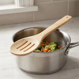 A light natural bamboo slotted spoon resting on a stainless steel saucepan filled with cooked broccoli and carrots, set on a white marble kitchen countertop.