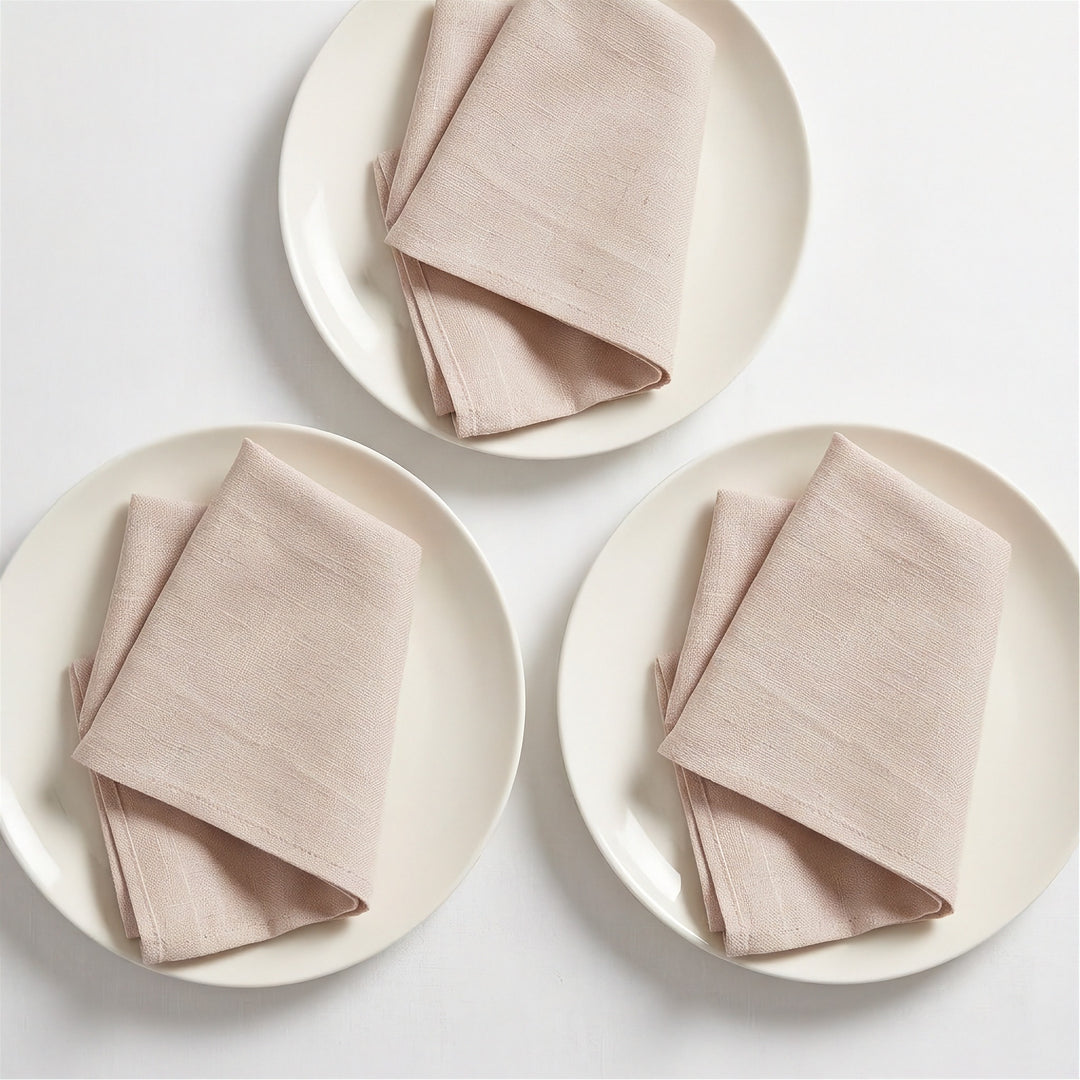 Three folded light blush pink linen napkins, each placed on a round off-white plate, arranged on a clean white background.