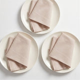 Three folded light blush pink linen napkins, each placed on a round off-white plate, arranged on a clean white background.