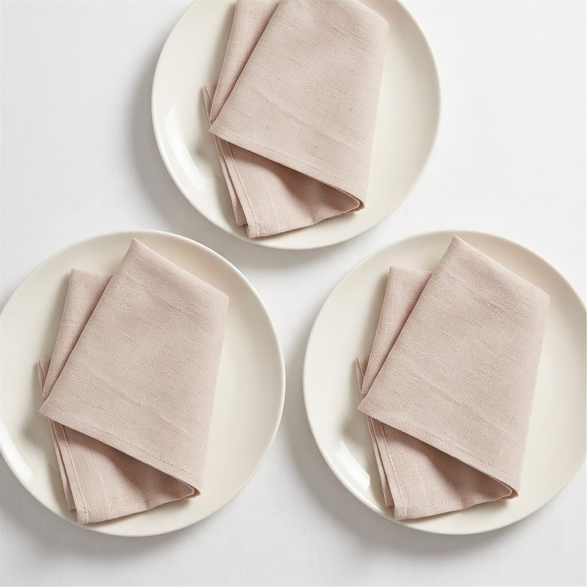 Three folded light blush pink linen napkins, each placed on a round off-white plate, arranged on a clean white background.