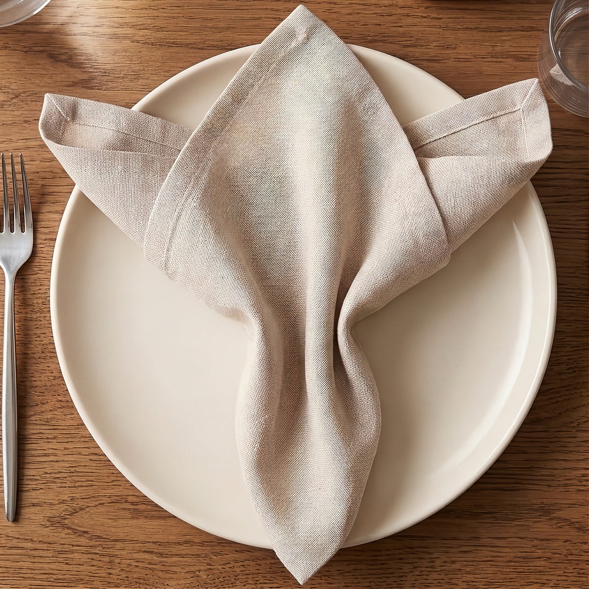 A folded beige linen napkin arranged on a round white plate, set on a warm brown wooden table with a silver fork partially visible on the left.