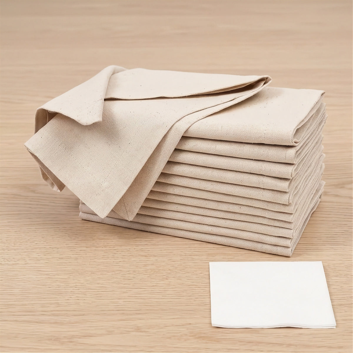 A stack of beige reusable cloth napkins on a light wood surface, with one white napkin placed in front.