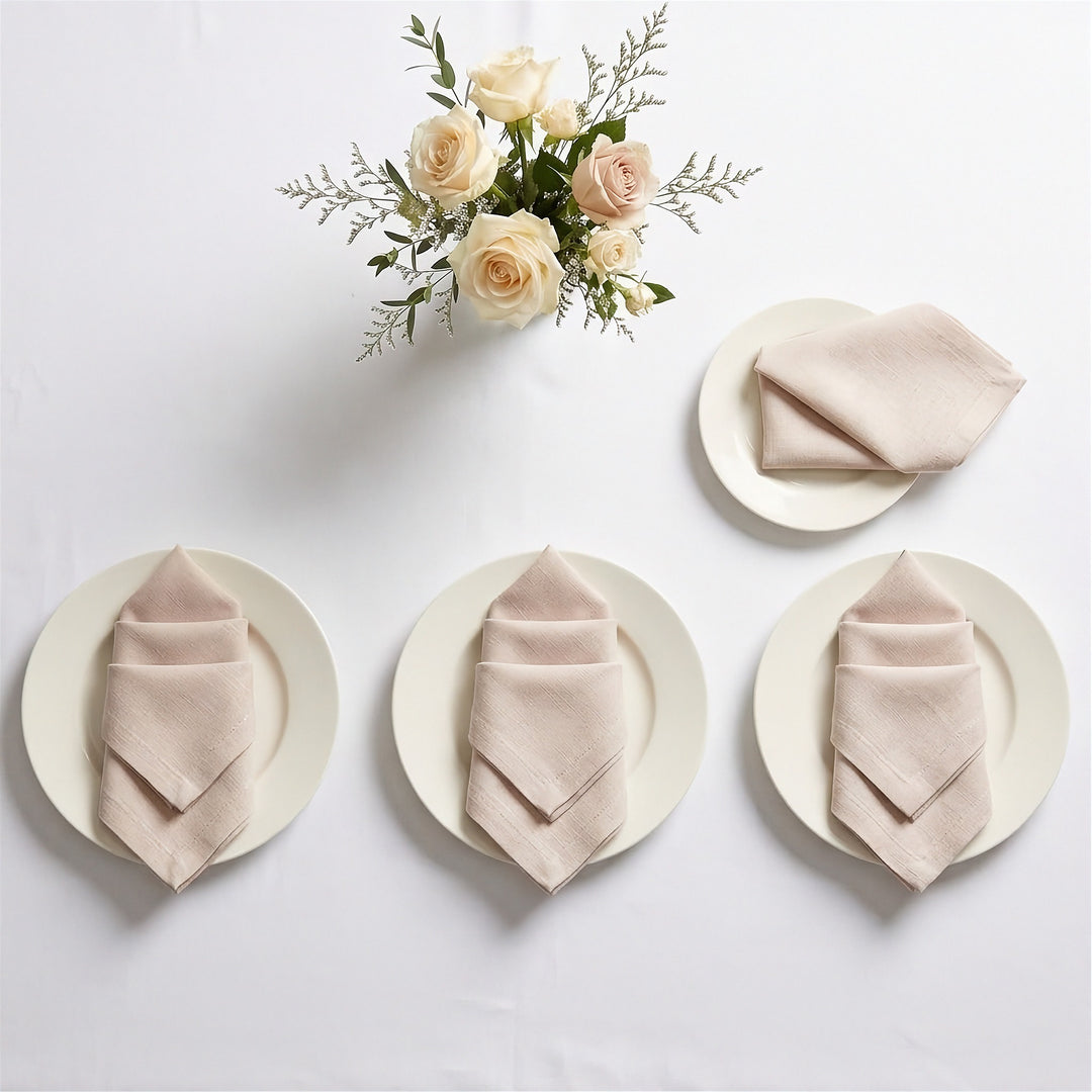 Three beige linen napkins, each folded and placed on a white plate, arranged on a white table with a small floral centerpiece above. No people or real environment are present, so this is Product/Packaging rather than Lifestyle.