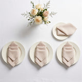 Three beige linen napkins, each folded and placed on a white plate, arranged on a white table with a small floral centerpiece above. No people or real environment are present, so this is Product/Packaging rather than Lifestyle.