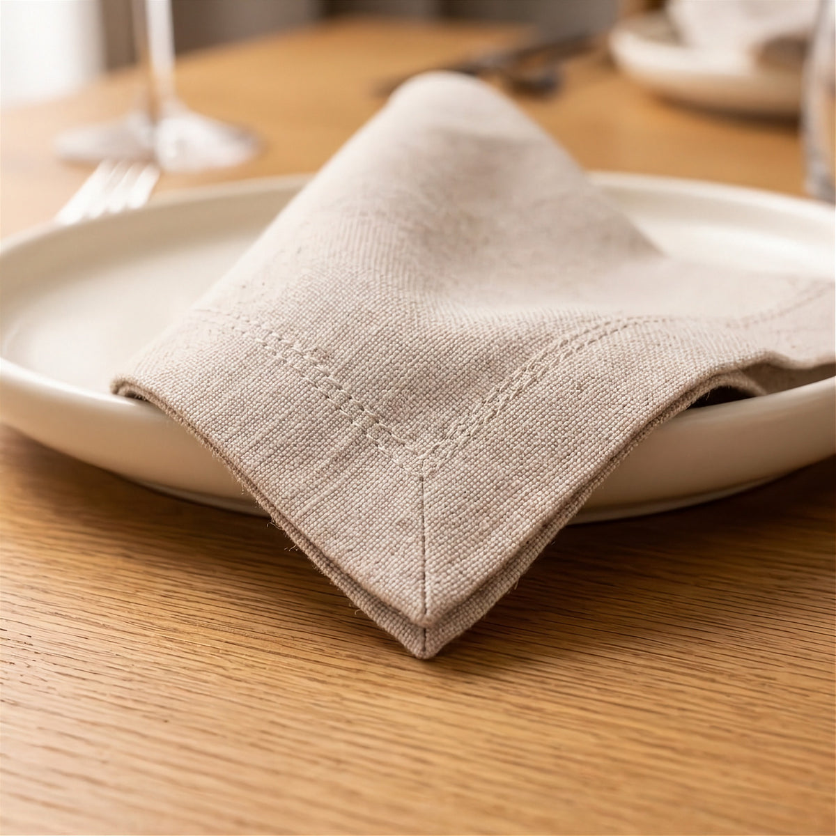 A folded beige linen napkin with decorative stitching rests on a white plate on a wooden table.