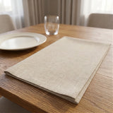 A folded beige linen placemat on a wooden dining table next to a white plate and a clear drinking glass.