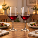 Two clear European-style red wine glasses with red wine on an oak dining table set for dinner