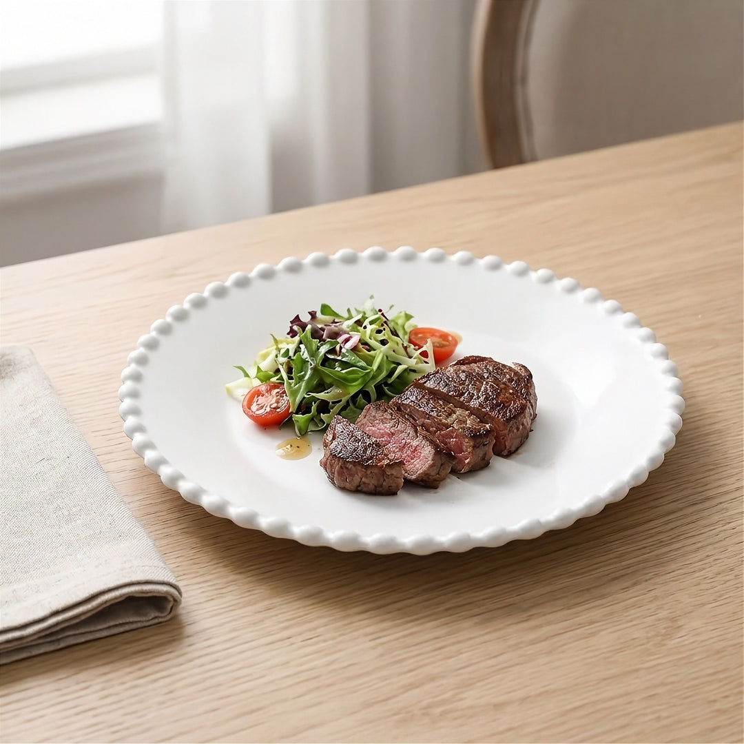 A white round plate with a beaded rim holds sliced steak and salad on a wooden table beside a beige napkin.