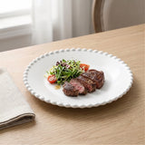 A white round plate with a beaded rim holds sliced steak and salad on a wooden table beside a beige napkin.