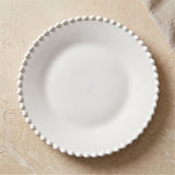 A white round plate with a raised beaded rim sits on a beige textured surface beside a fabric napkin.