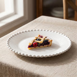 A white beaded-rim plate holds a slice of lattice berry tart on a beige tablecloth near a window.